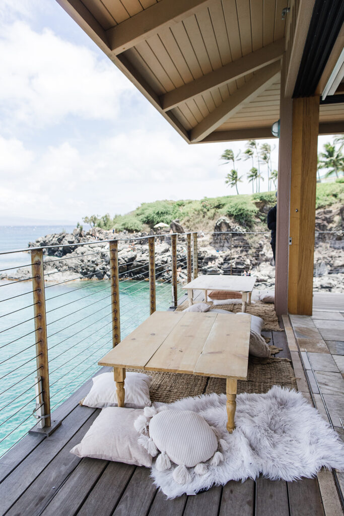 Matias Ezcurra Photography, Relaxing oceanfront seating area with wooden tables and fluffy pillows, perfect for a Maui wedding weekend guest event.