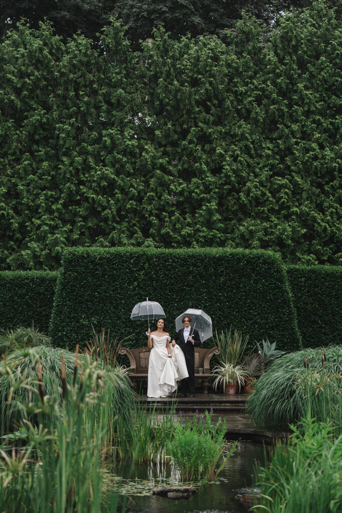Matias Ezcurra Photography, Bride and groom at Wave Hill in New York, sitting on a bench with umbrellas.
