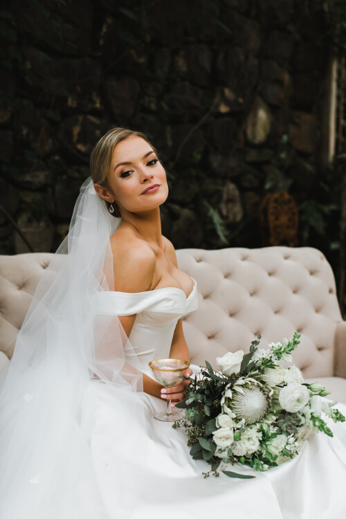 Matias Ezcurra Photography, Bride in off-the-shoulder gown sits on a couch, holding a champagne coupe and wedding bouquet at her Haiku Mill wedding.