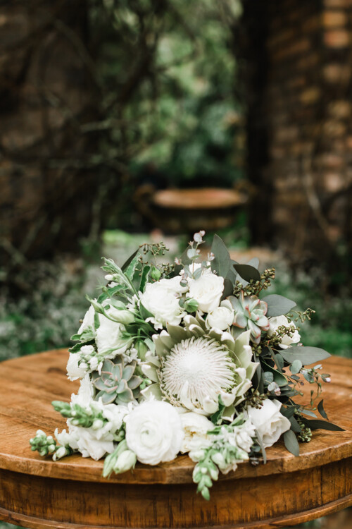 Matias Ezcurra Photography, Elegant white wedding bouquet featuring protea, roses, and succulents, resting on a wooden table.