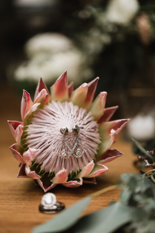 Matias Ezcurra Photography, Diamond earrings and rings rest on a pink protea flower at a Haiku Mill wedding.
