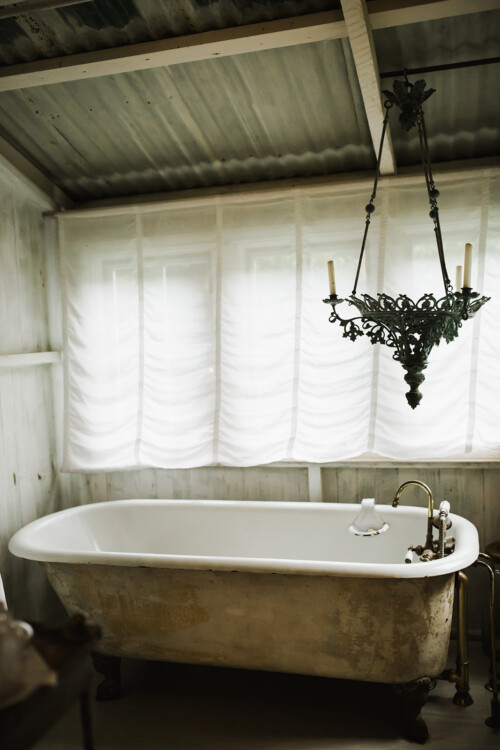 Matias Ezcurra Photography, Antique clawfoot tub in a rustic bathroom with a vintage chandelier.