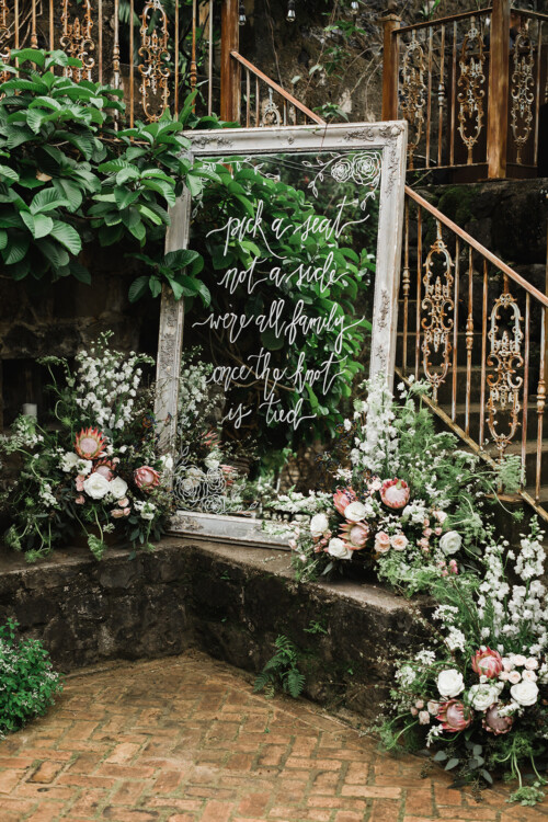 Matias Ezcurra Photography, Ornate mirror wedding sign: "Pick a seat, not a side, we're all family once the knot is tied." Floral arrangements flank the mirror.