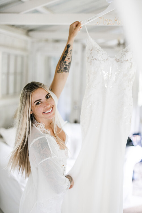 Matias Ezcurra Photography, Bride in white robe admires her wedding dress on a hanger at Haiku Mill.
