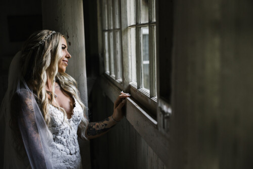 Matias Ezcurra Photography, Bride in lace wedding dress looks out window at Haiku Mill.