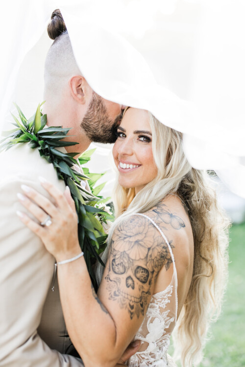 Matias Ezcurra Photography, Bride and groom embrace under a veil at their Haiku Mill wedding. The bride shows off a floral skull tattoo.