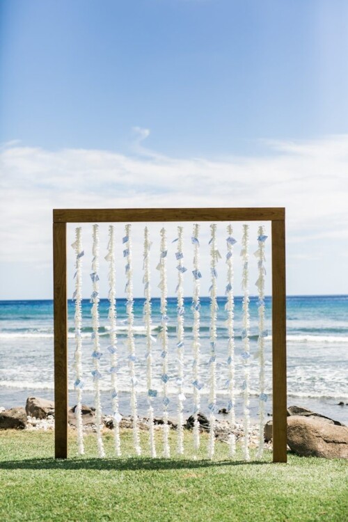 Matias Ezcurra Photography, Wooden wedding ceremony backdrop with white flower lei and blue place cards, ocean view.