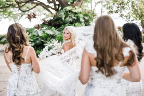 Matias Ezcurra Photography, Bride in lace wedding dress with flowing veil, smiling at bridesmaids in floral print dresses at Olowalu Plantation wedding.