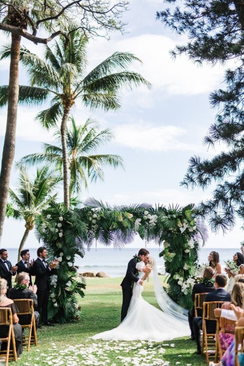 Matias Ezcurra Photography, Bride and groom kiss at their Olowalu Plantation wedding ceremony, under a lush tropical floral arch.