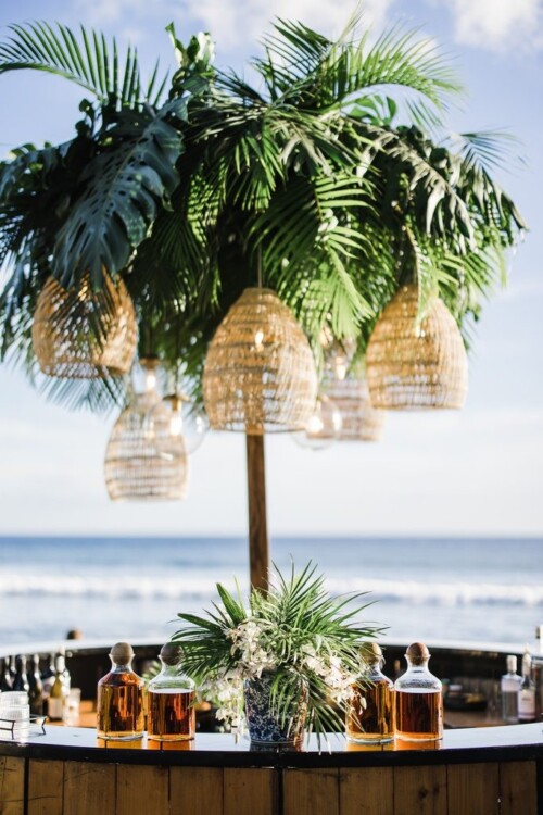Matias Ezcurra Photography, Beach wedding bar with tropical decor, wicker pendant lights, and bottles of liquor. Olowalu Plantation wedding.