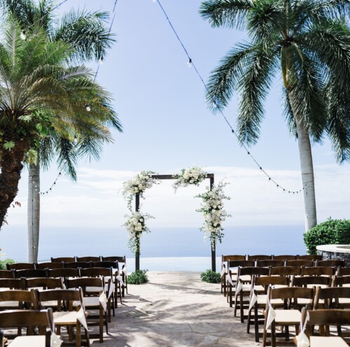 Matias Ezcurra Photography, Oceanfront wedding ceremony setup with white floral arch and wooden chairs.