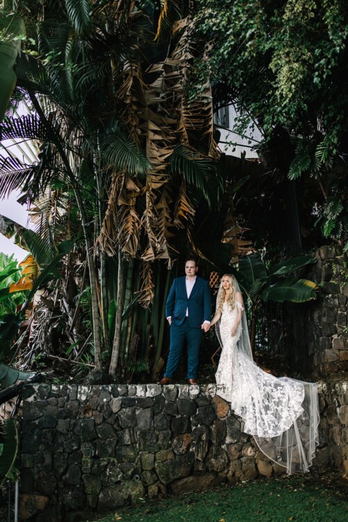 Matias Ezcurra Photography, Bride and groom holding hands, posing on a stone wall amidst lush tropical foliage.