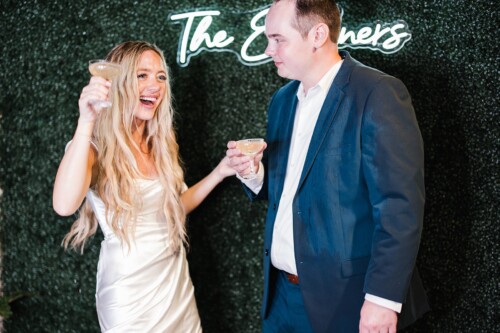 Matias Ezcurra Photography, Happy bride and groom toasting with champagne, "The Ewers" neon sign in background.
