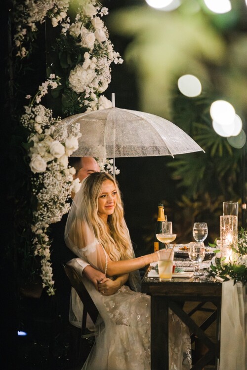 Matias Ezcurra Photography, Bride and groom seated under a clear umbrella at their wedding reception.