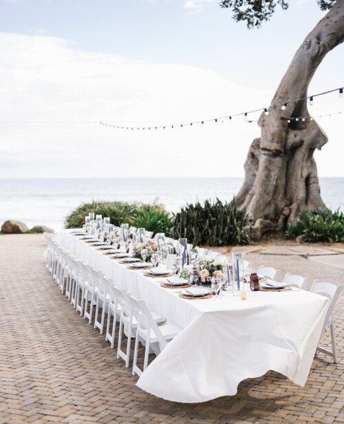 Matias Ezcurra Photography, Long wedding reception table with white linens, ocean view, and string lights.