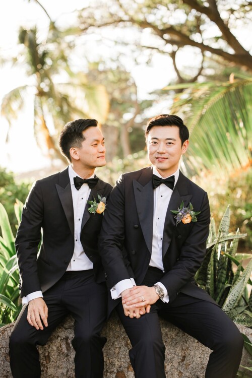 Matias Ezcurra Photography, Two grooms in black tuxedos hold hands, smiling at each other in a tropical garden.