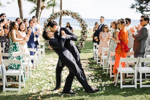 Matias Ezcurra Photography, Two grooms share a passionate kiss at their beach wedding ceremony, surrounded by cheering guests.