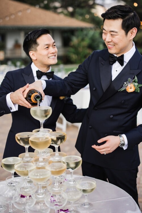 Matias Ezcurra Photography, Two grooms in tuxedos pour champagne into a wedding champagne tower.
