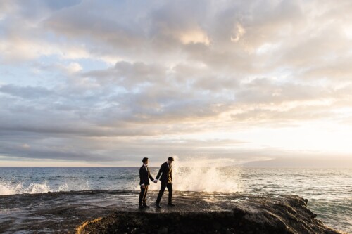 Matias Ezcurra Photography, Two grooms in tuxedos hold hands on a rocky ocean shore as waves crash.