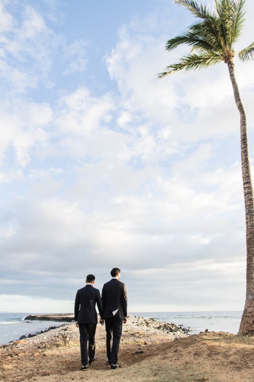 Matias Ezcurra Photography, Two grooms in black suits hold hands, walking away from the camera toward the ocean under a cloudy sky and a palm tree.