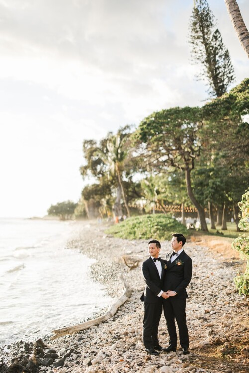 Matias Ezcurra Photography, Two grooms in black tuxedos hold hands on a beach at sunset.