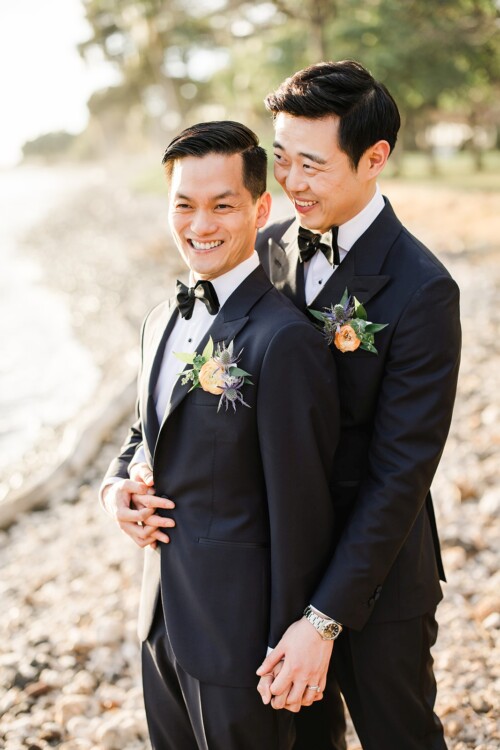 Matias Ezcurra Photography, Two happy grooms in black tuxedos hold hands on a beach.
