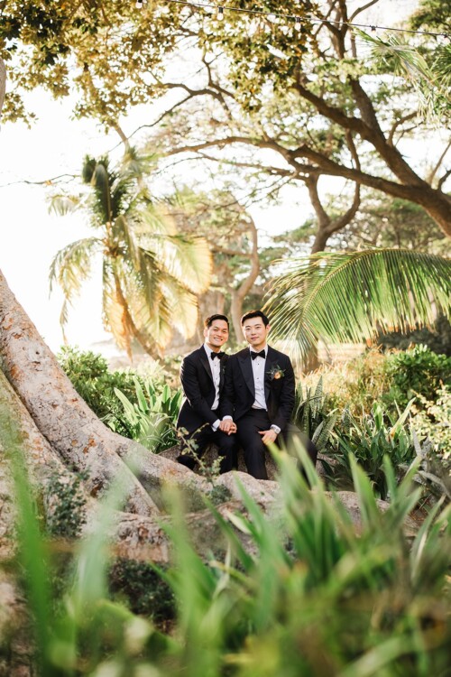 Matias Ezcurra Photography, Two grooms in tuxedos smiling, sitting on tree roots in a lush garden.
