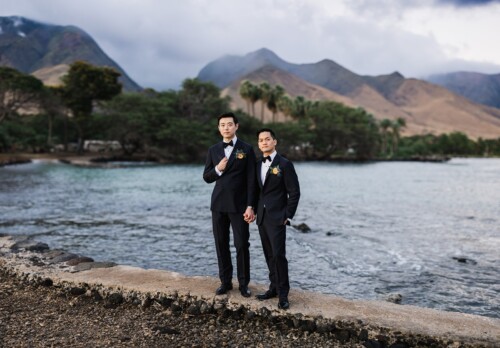 Matias Ezcurra Photography, Two grooms in tuxedos holding hands on a seaside dock, mountains in the background.