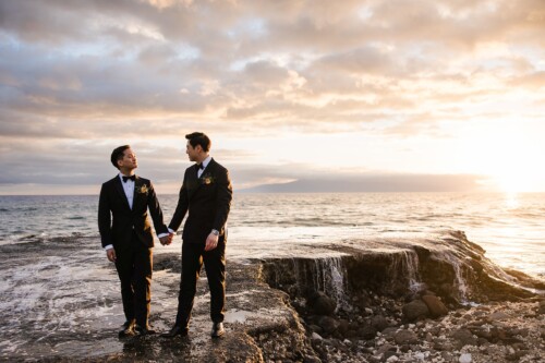 Matias Ezcurra Photography, Two grooms in black tuxedos hold hands on a rocky ocean shore at sunset.