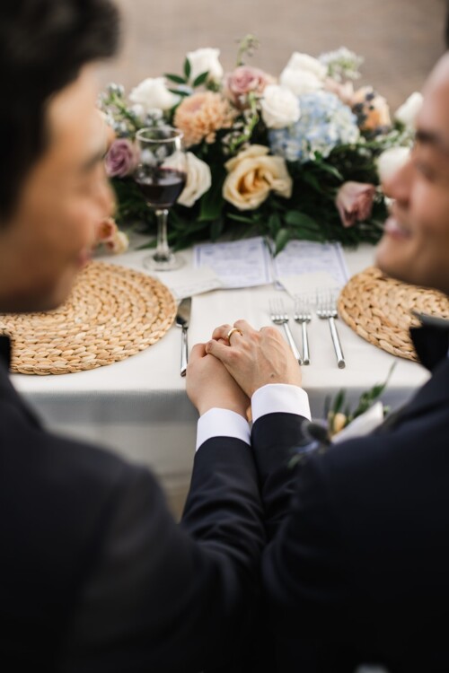 Matias Ezcurra Photography, Two grooms hold hands at their wedding reception, seated at a table with a floral centerpiece.