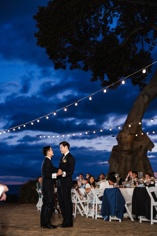 Matias Ezcurra Photography, Gay couple's first dance at sunset wedding reception under string lights.