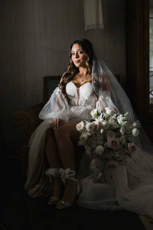 Matias Ezcurra Photography, Bride in off-the-shoulder wedding dress sits on a chair with bouquet at Olowalu Plantation House.
