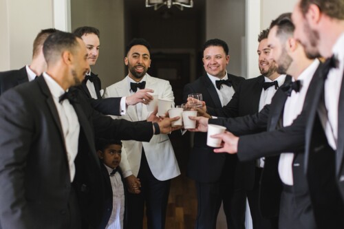 Matias Ezcurra Photography, Groomsmen in tuxedos toast with drinks before a Kualoa Ranch wedding.