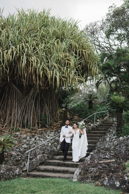Matias Ezcurra Photography, Bride and groom in white wedding attire descend stone steps at Kualoa Ranch.
