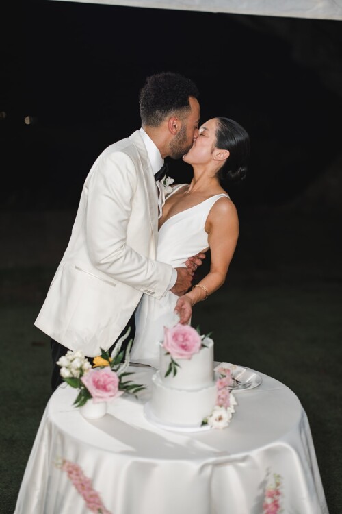 Matias Ezcurra Photography, Bride and groom kiss after cutting their elegant wedding cake at their Kualoa Ranch wedding.