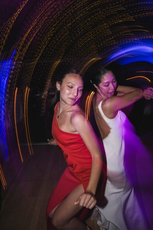 Matias Ezcurra Photography, Two women dance at a Kualoa Ranch wedding reception, illuminated by colorful lights.