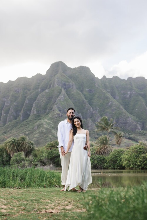 Matias Ezcurra Photography, Klaire & Alex hold hands, smiling at Kualoa Ranch, Hawaii, in front of mountains.