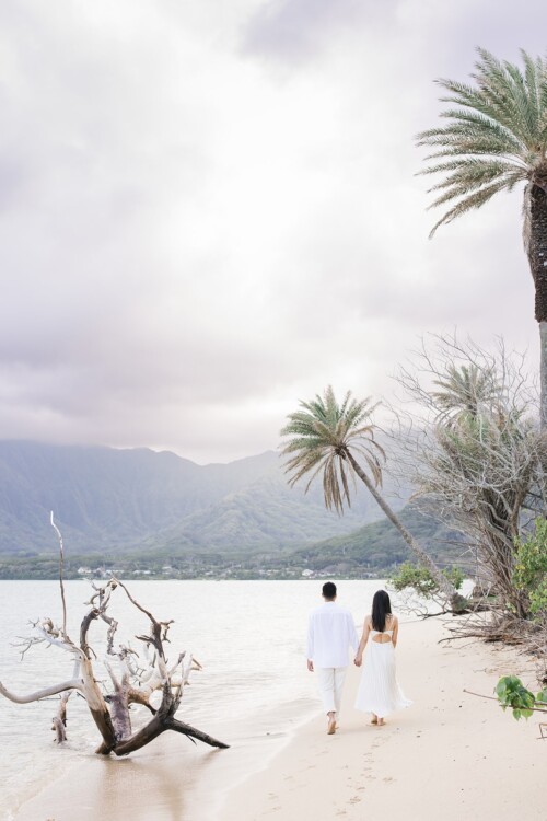 Matias Ezcurra Photography, Couple in white attire holding hands, walking on a beach at Kualoa Ranch.