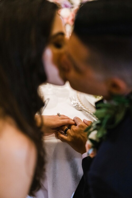 Matias Ezcurra Photography, Newlyweds' hands clasped, showcasing wedding rings; a tender kiss in the background.